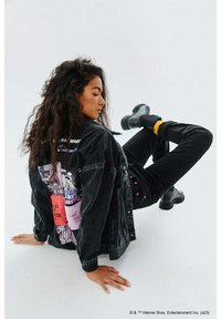 Czarna kurtka dżinsowa z kolorowymi nadrukami z tyłu, noszona z dopasowanymi czarnymi spodniami i czarnymi botkami. Prosty, nowoczesny design.