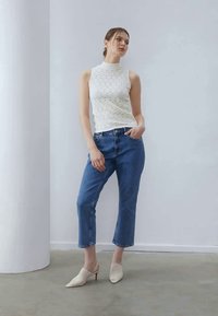 Strikket hvid ærmeløs top med høj hals, parret med blå opsmøgede jeans og cremefarvede åbne mule-sko på et glat gråt gulv mod en enkel baggrund.