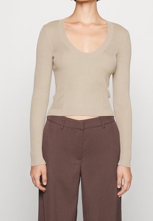 Beige ribbet langærmet top med dyb scooped halsudskæring kombineret med højtaljede, vide brune bukser på en stående person.