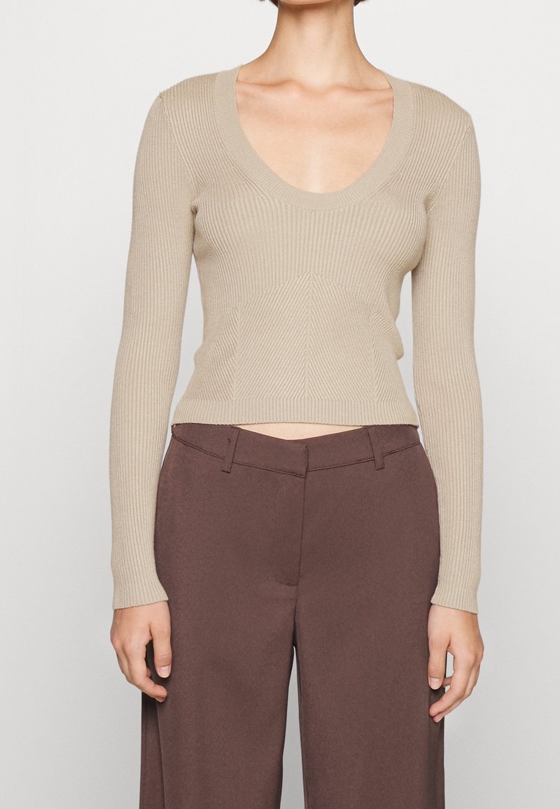 Haut beige côtelé à manches longues avec un col rond profond, associé à un pantalon marron taille haute et jambes larges sur une personne debout.