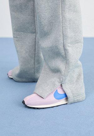 Person iført løstsiddende grå joggingbukser, der dækker pink sneakers med blå Nike swoosh, stående på lyst blåt tæppe.