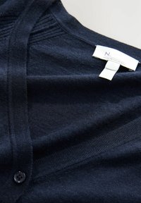 Navyblauer Cardigan aus weichem Stoff, mit V-Ausschnitt, vertikalem Rippenmuster und einem einzigen schwarzen Knopfverschluss. Etikett sichtbar.