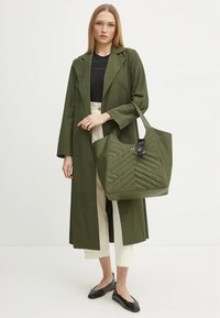 Bolso tote acolchado de color verde oliva con forma estructurada y herrajes negros en contraste. La modelo lleva un abrigo largo a juego y unos zapatos planos negros.