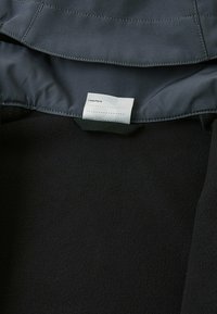 Intérieur d'une veste en polaire noire avec une étiquette affichant "J'étais ici." et des espaces vides pour des informations. La texture douce du tissu est visible.