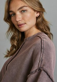 Felpa oversize in morbido tessuto mauve, con colletto a coste e orli sfrangiati. Capelli acconciati in onde morbide.