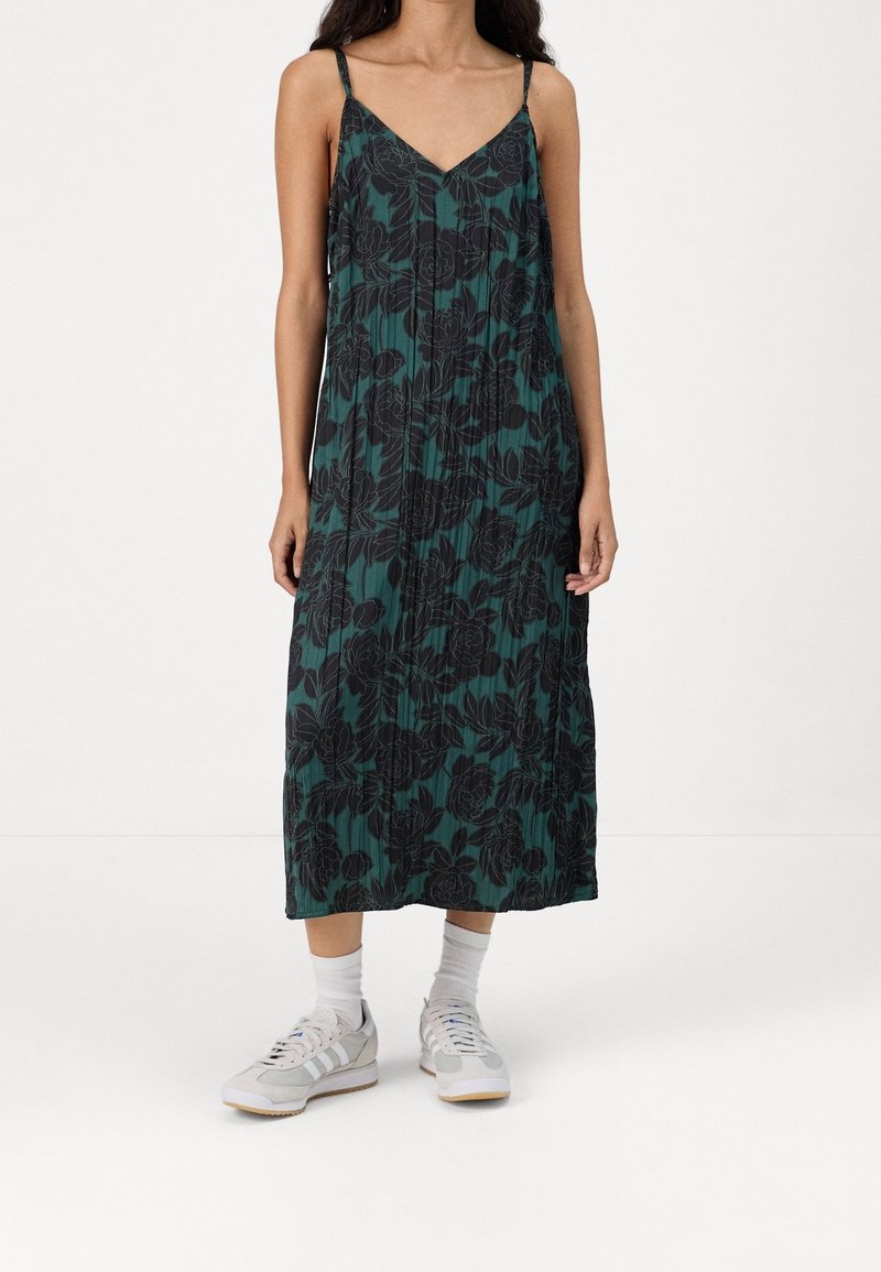 Femme portant une robe midi fleurie vert foncé à fines bretelles, associée à des chaussettes blanches et des baskets gris clair.