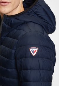 Marinblå puffjacka med horisontellt kviltat mönster, med huva och en synlig Rossignol-logopatch på ärmen. Slät, glänsande tyg.