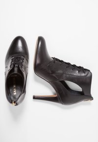 Mustat nahkaiset korkokengät, joissa on nyöritys, terävä kärki ja sivuilla leikkaukset. Varustettu kullanvärisellä vetoketjulla ja teksturoiduilla paneelidetaljeilla.