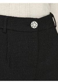 Černé texturované kalhoty mají dekorativní knoflík ve tvaru květiny s bílými okvětními lístky a centrálním drahokamem, což dodává jemný akcent.