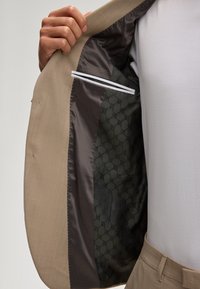 Linke Hand hält eine offene beige Anzugjacke, die ein dunkles gemustertes Futter und eine Innentasche zeigt, getragen mit weißem Hemd und passenden beigen Hosen.