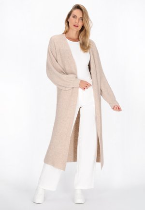 Langer, beiger Cardigan aus strukturiertem Strickmaterial mit offener Vorderseite, überschnittenen Schultern und weiten Ärmeln, kombiniert mit weißen Hosen und Sneakers.