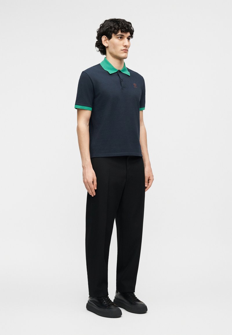 Man staat en draagt een marineblauw poloshirt met groene kraag en mouwboorden, zwarte broek en zwarte sneakers tegen een effen achtergrond.