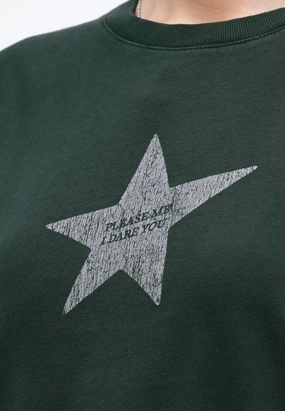 Hanorac de culoare verde închis cu o mare stea argintie uzată, având inscripția "ÎNDEMNĂ-MĂ, ÎNDRĂZNEȘTE" centrată în interiorul stelei.