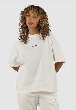 Nainen, jolla on kiharat hiukset ja yllä löysä valkoinen t-paita sekä siihen sopivat valkoiset housut, joissa molemmissa lukee "PEGADOR".