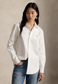Kvinde med langt mørkt hår iført hvid skjorte med knapper og blå jeans, stående med den ene hånd i lommen mod en neutral baggrund.