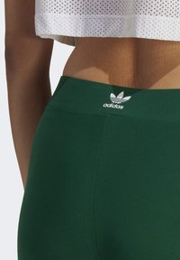 Gröna träningsleggings med en slät textur och hög midja, med en vit Adidas-logga på baksidan. Crop topen har ett perforerat mönster.