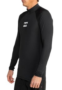 Hombre vistiendo una parte superior de traje de neopreno negro de manga larga con un pequeño logo de ola blanca en el pecho, de pie contra un fondo blanco.