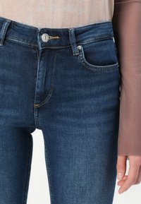 Donkerblauwe skinny jeans hebben een soepele textuur, voorzakken en een metalen knoopsluiting in de taille met lichte stikselaccenten.