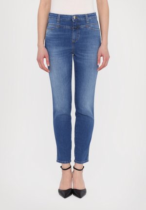 Femme portant un jean skinny bleu taille mi-haute et des talons noirs à bride cheville, debout devant un fond blanc uni.