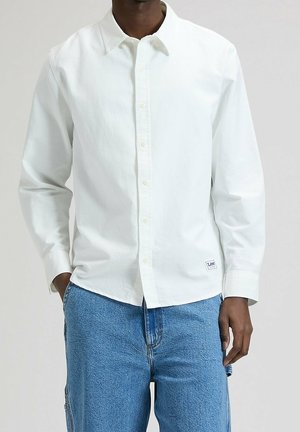 Mand iført en hvid skjorte med lange ærmer og blå denimjeans med hænder hvilende nær lommer mod en ensfarvet baggrund.