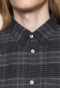 Gros plan sur une personne portant une chemise à carreaux gris foncé avec des boutons blancs et un col plié.