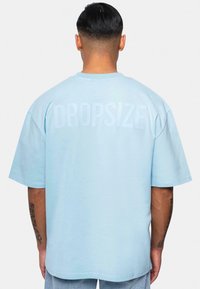 Lyseblå oversized t-shirt med korte ærmer, der har teksten "DROPSIZE" i et subtilt, præget design på ryggen. Blødt bomuldsstof.