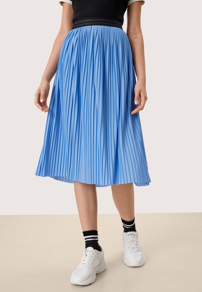 Donna che indossa una gonna midi plissettata blu, un top nero a maniche corte, calze nere con strisce bianche e sneakers bianche, in piedi su un pavimento beige.