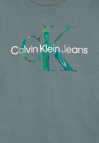 Tricou gri cu inscripția "Calvin Klein Jeans" în alb, având literele îndrăznețe "CK" care prezintă un model marmorat verde și albastru în interior.
