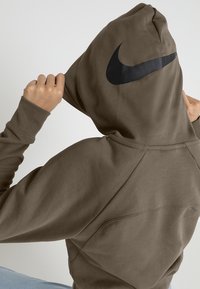 Brun Nike-hoodie med en stor svart logga på ryggen, med ett texturerat tyg och en avslappnad passform. Huvtröja med långa ärmar.