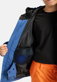Personne tenant une veste bleue ouverte pour montrer la poche intérieure en maille noire, portant un t-shirt noir et un pantalon orange sur fond blanc.