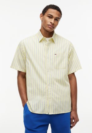 Junger Mann trägt ein gelbes kurzärmeliges Hemd mit blauen vertikalen Streifen und blaue Hosen, steht mit einer Hand in der Tasche.