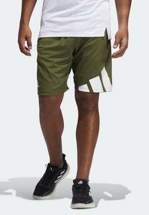 Homme portant un short vert olive avec un motif blanc, une chemise blanche et des baskets noires Adidas, debout sur un fond clair.