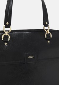 Sac à main en cuir noir avec quincaillerie dorée, double poignées supérieures et détail de logo embossé. Texture lisse avec un motif de grain subtil.