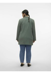 Manteau long vert olive avec une coupe droite, présentant un col plat et une texture lisse. Associé avec un jean bleu et des chaussures noires plates.