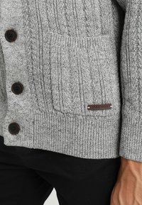 Grå strikket cardigan med ribbestruktur, fire brune knapper, en frontlomme og et lite lærmerke som sier "PIER-ONE."