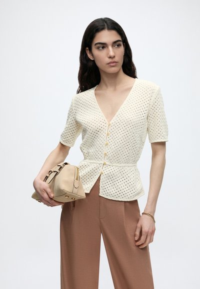 Γυναίκα με μακριά σκούρα μαλλιά φοράει ένα εκρού πλεκτό τοπ με κουμπιά και καφέ παντελόνι, κρατώντας μια μπεζ τσάντα με χρυσά εξαρτήματα στο αριστερό της χέρι.