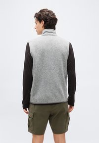 Άνδρας γυρισμένος πίσω φορά γκρι αμάνικο φλις γιλέκο πάνω από μαύρο μακρυμάνικο πουκάμισο και χακί cargo σορτς πάνω σε απλό φόντο.