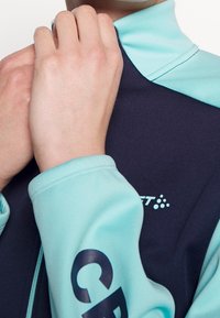 Chaqueta deportiva con cremallera en azul marino y verde azulado, que cuenta con una textura suave, un alto cuello y un logotipo impreso en la manga. Primer plano de manos ajustando el cuello.