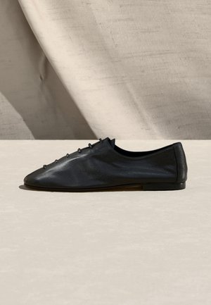 Chaussure plate en cuir noir avec bout pointu et petits œillets en boucle, vue de côté sur une surface claire avec un fond en tissu beige.