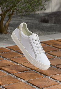 Baltas Tommy Hilfiger sportinis batelis su auksiniais akcentais ir raišteliais, išdėstytas ant medinių kvadratinių plytelių ir mažų pilkų akmenėlių lauke.