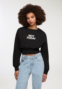 Giovane donna con capelli ricci e voluminosi che indossa un felpa nera corta con la scritta "NOT TODAY" e jeans chiari a vita alta.