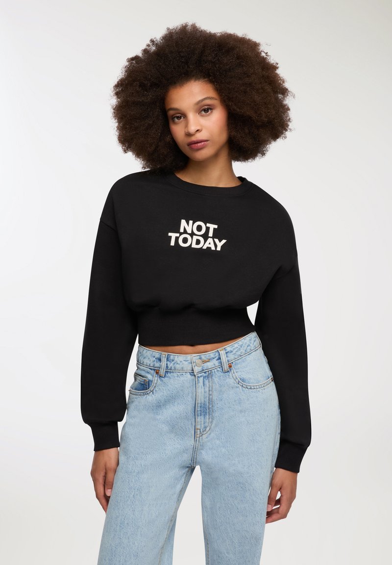 Giovane donna con capelli ricci e voluminosi che indossa un felpa nera corta con la scritta "NOT TODAY" e jeans chiari a vita alta.