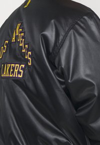 Chaqueta de satén negra con el texto bordado "Los Angeles Lakers" en púrpura y amarillo en la parte posterior del hombro.