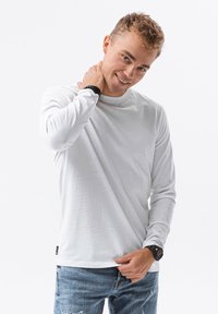 Weißes Longsleeve-T-Shirt mit Rundhalsausschnitt, kombiniert mit hellblauen, verwaschenen Jeans. Das Modell trägt eine schwarze Armbanduhr.