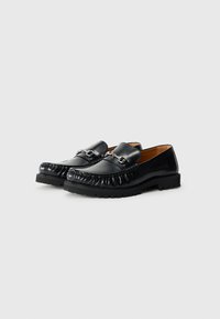 Mocasines de cuero negro con punta redondeada, que presentan un diseño trenzado y acentos de hardware en tono plateado en el empeine. Suela texturizada.