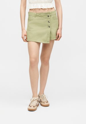 Femme portant une mini-jupe boutonnée vert clair, un top court blanc et des baskets beige avec des taches noires, debout devant un fond uni.