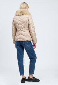 Beige puffermantel med pelskrave, chevron-quiltet design og vandrette sømme. Bæres med høj midje blå jeans og sorte loafers.