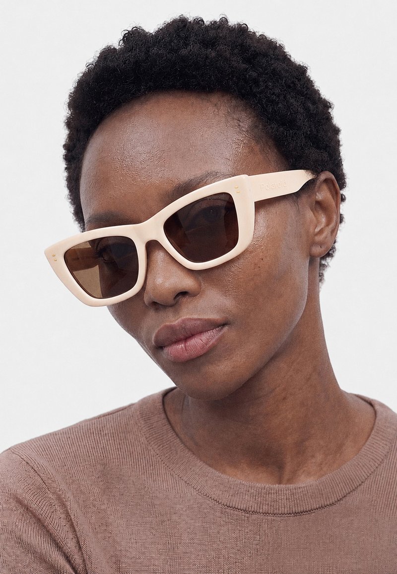 Personne aux cheveux courts et bouclés portant des lunettes de soleil rectangulaires beiges et un haut en maille marron clair sur un fond uni.