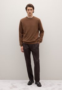 Pull en laine marron avec un col rond, associé à un pantalon plissé marron foncé. Le modèle porte des mocassins noirs. Fond neutre.
