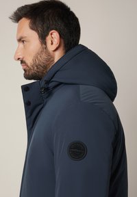 Mężczyzna z ciemną brodą, noszący granatową kurtkę z kapturem i okrągłą czarną łatą na lewym rękawie, zwrócony w lewo na neutralnym tle.